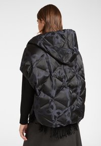 Kobieta z brązowymi włosami ubrana w czarny pikowany płaszcz z kapturem, zdobiony okrągłymi wzorami i czarnym frędzlami u dołu, pokazana od tyłu.