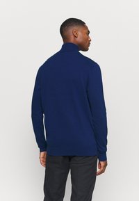 Marineblauer Pullover mit hohem Kragen, gerippten Bündchen und Saum, aus einem glatten Strickmaterial gefertigt, von hinten an einem Modell mit dunklen Hosen gezeigt.