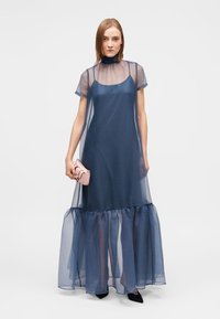 Μακρύ, διαφανές φόρεμα σε ναυτικό μπλε χρώμα με κοντά μανίκια, τελείωμα με βολάν και υφή. Συνδυασμένο με μικρό ροζ τσάντα χειρός και μαύρα παπούτσια.