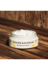 Bocal en verre transparent de beurre corporel avec une texture crème, présentant un motif d'oiseau jaune et un texte noir sur une étiquette blanche.