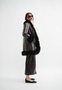 Bruine faux lederen jas met zwarte faux bontafwerking. Lange mouwen, zijzakken, gecombineerd met een lange bruin rok en zwarte flats.