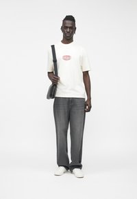 Camiseta blanca con el logo rojo "GUESS", vaqueros grises de corte holgado y zapatillas blancas. Lleva un bolso mensajero negro pequeño.