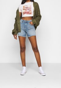 Olivi vihreä ylipitkä neulecardigan valkoisen graafisen printtipaidan päällä, yhdistettynä vaaleansinihin farkkushortseihin ja valkoisiin korkeavartisiin tennareihin.