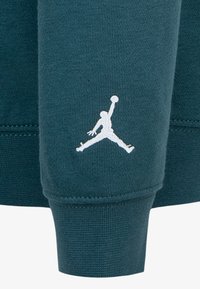 Tamsiai žalia džemperio medžiaga su gipiūriniais rankogaliais, kairiajame rankovėje esantis baltas siuvinėtas Jumpman logotipas.