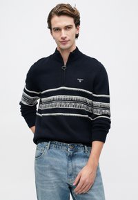 Młody mężczyzna w granatowym swetrze z białymi wzorzystymi paskami i pół-zamkiem, w połączeniu z jasnoniebieskimi dżinsami, stojący na jednolitym tle.