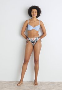 Lys blå bh med underwire, kombinert med høydejusterte svarte bukser med blomstertrykk og sidesnøring. Glatt stoff og tilpasset passform.