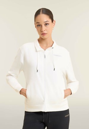 Jeune femme aux cheveux tirés en arrière, portant un sweat à capuche blanc zippé et un pantalon noir, les mains dans les poches, debout devant un fond clair uni.