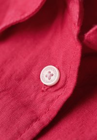 Nabij een rode stof met een witte knop waarop de tekst "SUPERDRY" en een bloemenpatroon staat. De textuur is glad en lichtjes geprofileerd.