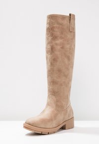 Beżowy kozaki do kolan z eko skóry o teksturowanym wykończeniu, zaokrąglonym nosku i masywnej podeszwie. Wyposażone w pętlę u góry ułatwiającą zakładanie.