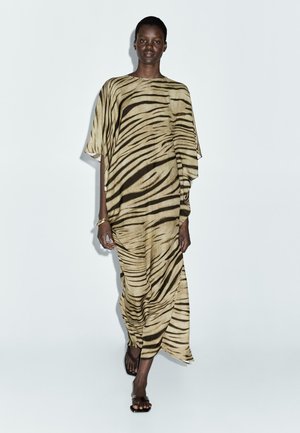 Robe longue fluide à imprimé animalier beige et marron. Tissu doux, manches larges et design drapé. Portée avec des sandales noires simples.