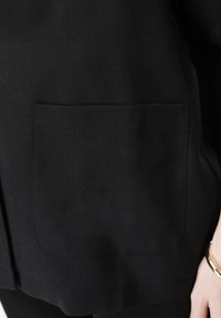 Detailní záběr černé látky s obdélníkovou náprsní kapsou a rukou na boku, která má na zápěstí zlatý náramek.