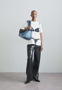 T-shirt bianca con grafica di reggiseno nero, abbinata a pantaloni in pelle neri a gamba larga. Il modello porta una borsa in denim blu, in piedi su uno sfondo neutro.