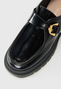 Black leather loafer with chunky sole, gold buckle strap, and rounded toe, shown from above on a white background.