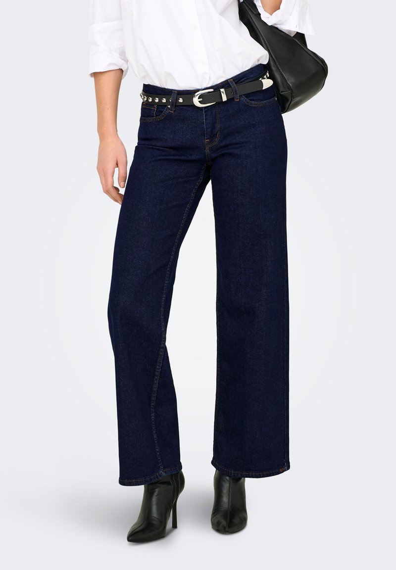 Jean large bleu foncé à taille haute, avec des accessoires argentés sur une ceinture noire, accompagné d'un sac à main noir et de talons noirs.