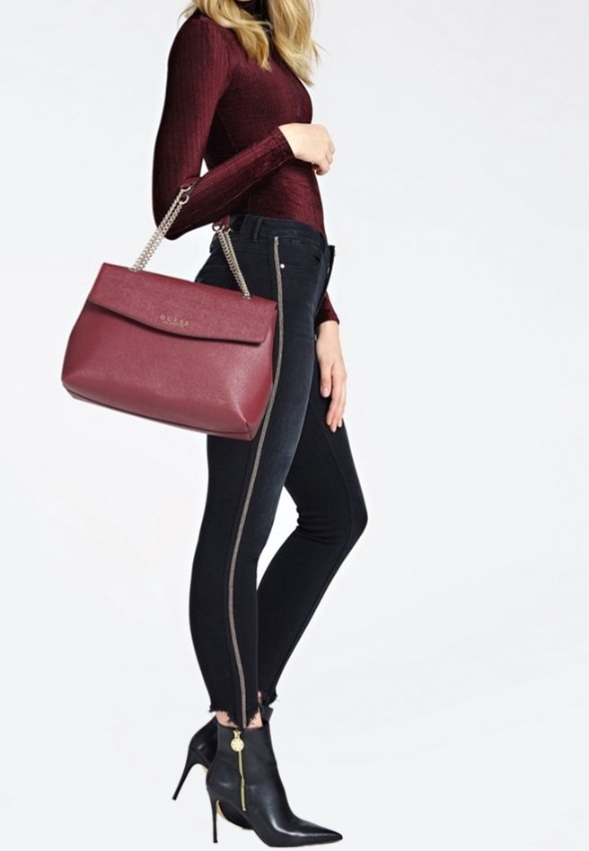 Femme portant un haut bordeaux et un jean skinny noir avec un détail de fermeture éclair, tenant un sac à main bordeaux assorti et portant des bottes à talons noires.