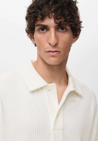 Polo blanco con canalé, cuello plegable y tapeta de tres botones. El material parece suave con una superficie texturizada.