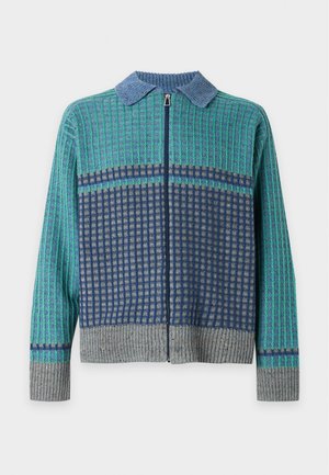 Jeune homme debout portant un pull zippé vert et bleu, un pantalon noir et des chaussures jaunes sur un fond blanc uni.