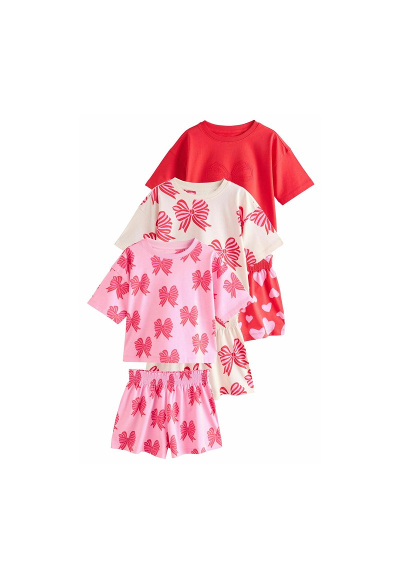 Tre set di abbigliamento per bambini con t-shirt a maniche corte e pantaloni corti. I colori includono rosso, rosa e crema con motivi a fiocco e cuori.