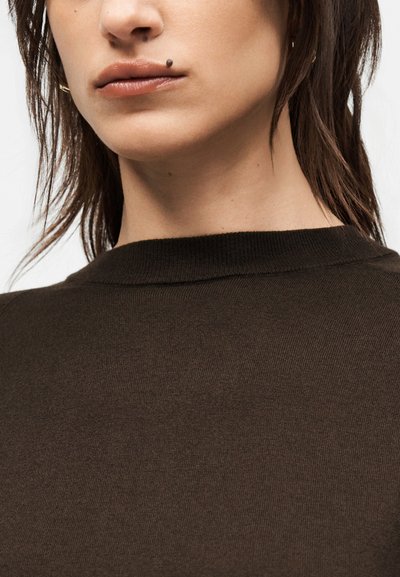 Gros plan sur la partie inférieure du visage et le cou d’une personne portant un pull marron foncé, avec une tâche de beauté bien visible au-dessus de la lèvre supérieure.