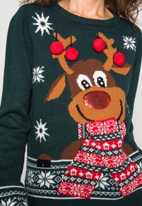 Pull vert en tricot présentant un motif de renne, avec des pompons rouges et des flocons de neige. Détails remarquables : une écharpe à motifs et un nez texturé.