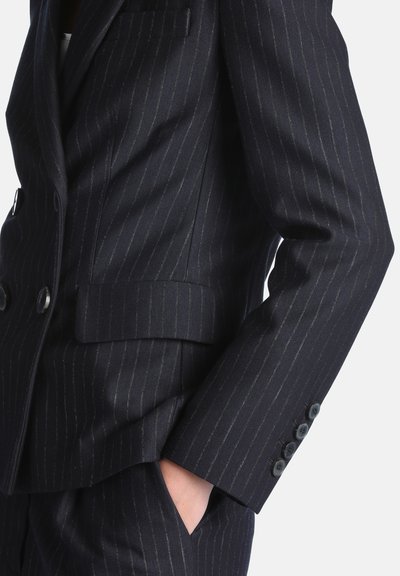 Veste de costume à fines rayures foncées et pantalon assorti avec une main dans la poche, montrant les détails des boutons sur la manche et le rabat de poche de la veste.