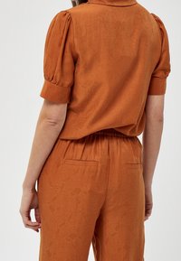 Rust-colored, short-sleeved blouse with a subtle textured pattern, featuring a relaxed fit and an elastic waistband. Back view shows a smooth finish.