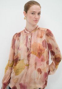Durchsichtige rosa Bluse mit einem Blattdruck in Gelb, Orange und Burgunderrot. Mit Knopfverschluss sowie Rüsche am Halsauschnitt und an den Ärmelabschlüssen.