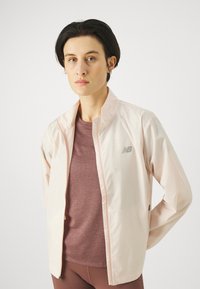 Giacca rosa chiaro con zip e colletto alto, con un logo sottile. Indossata sopra un top bordeaux lavorato. Materiale liscio con una vestibilità aderente.