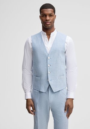 Mann trägt hellblaues Weste und passende Hose über weißem Hemd, steht vor einfachem grauem Hintergrund.
