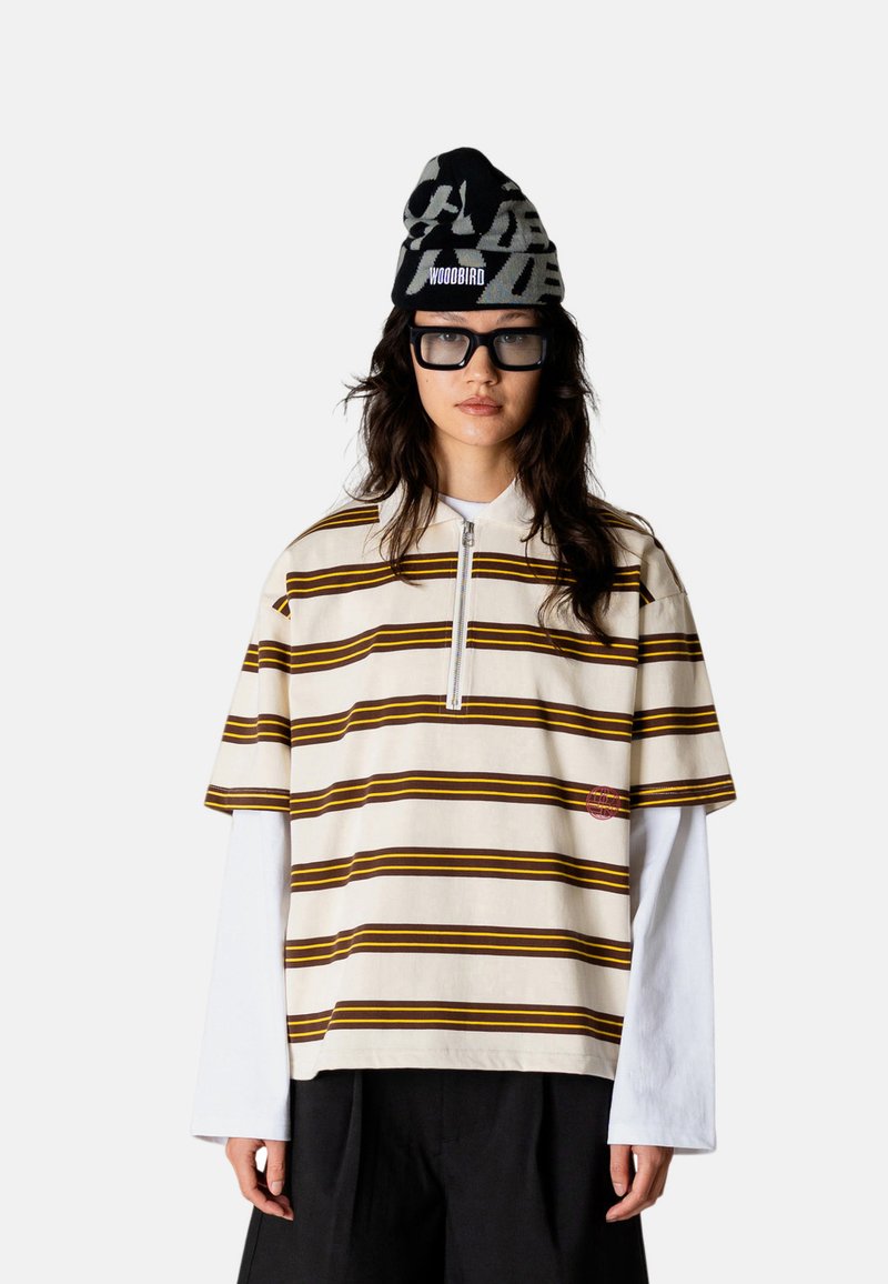 Femme portant une chemise rayée à manches courtes oversize sur des manches longues blanches, un pantalon noir à jambes larges, un bonnet noir avec texte et de grandes lunettes noires.