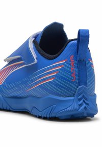 Zapatilla deportiva azul con un material sintético liso y texturizado; cuenta con una correa de Velcro y patrones angulares en un azul más claro y acentos naranjas.