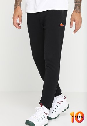 Homme portant un pantalon noir slim-fit avec un logo sur la cuisse et des baskets blanches avec des semelles vertes et des touches rouges sur fond uni.