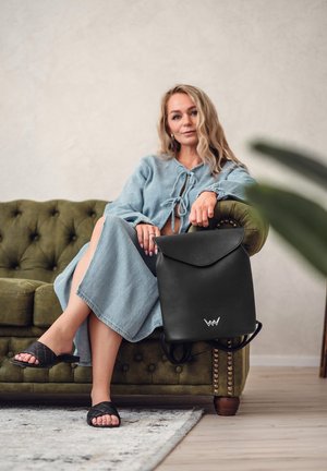 Schwarzer Leder-Rucksack mit Klappdesign und Logo, neben einer Person auf einem grünen, gesteppte Sofa platziert. Der Hintergrund ist neutral.