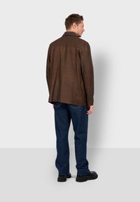 Chaqueta de cuero marrón con cuello, costuras en los hombros y dos botones en la parte posterior. Llevada sobre vaqueros azules y zapatos negros.