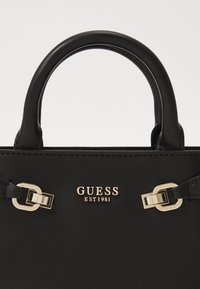 Black handbag made of smooth material, featuring a rounded handle, gold hardware, and the brand name "GUESS EST 1981" embossed in front.