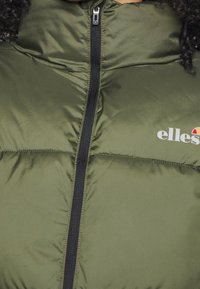 Giacca puffer verde oliva con zip nera, caratterizzata da un motivo a trapunta e un logo sul lato sinistro, realizzata in un materiale liscio e lucido.