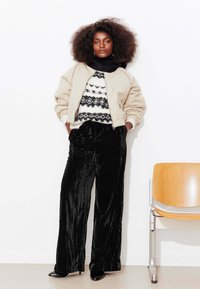 Veste bomber beige portée sur un pull à motifs noir et blanc, associée à un pantalon large en velours noir et des talons pointus noirs.