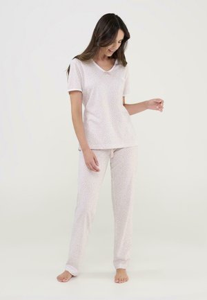 Mujer de pie descalza, vistiendo una blusa de manga corta color rosa claro y pantalones largos a juego con un sutil estampado de leopardo sobre un fondo liso.