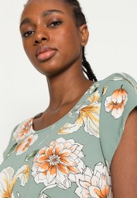 Top floral com fundo verde e padrões de flores laranja e brancas. As mangas curtas e o decote redondo aprimoram o seu design.