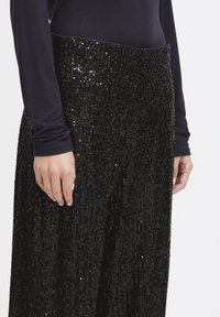 Pantalon noir à sequins avec une large ceinture et une coupe ample. La texture est brillante, avec un dessus lisse et des sequins scintillants sur toute la surface.