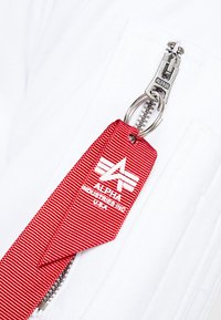 Etichetta in tessuto rosso con righe verticali, cerniera argentata etichettata "ALPHA," attaccata a un indumento bianco. Presenta un logo con tre righe e testo.