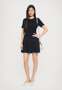 Tengerészkék, rövid ujjú ruha, bordázott textúrával a felsőrészen, bővülő szoknyával és függőleges csíkokkal. Világos színű táskával párosítva.