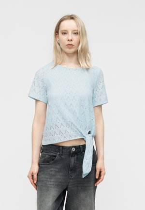 Kvinde iført en lyseblå bluse med korte ærmer og hulmønster, bundet i taljen, samt falmede sorte jeans, stående mod en ensfarvet lys baggrund.