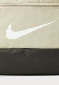 Sac en tissu beige avec une surface texturée ; comporte un grand logo Swoosh blanc de Nike et une section inférieure noire unie. Fermeture zippée en haut.