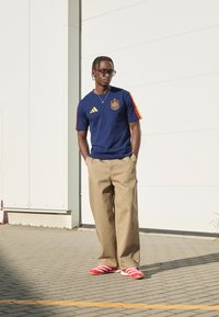 Maglietta Adidas blu navy con strisce rosse e gialle, pantaloni larghi color khaki e scarpe rosse con dettagli bianchi. La persona indossa occhiali e gioielli in argento.