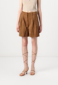Shorts plissés en daim marron avec une taille mi-haute, associés à des sandales à talons hauts beige avec lanières ; texture lisse et coupe ajustée.