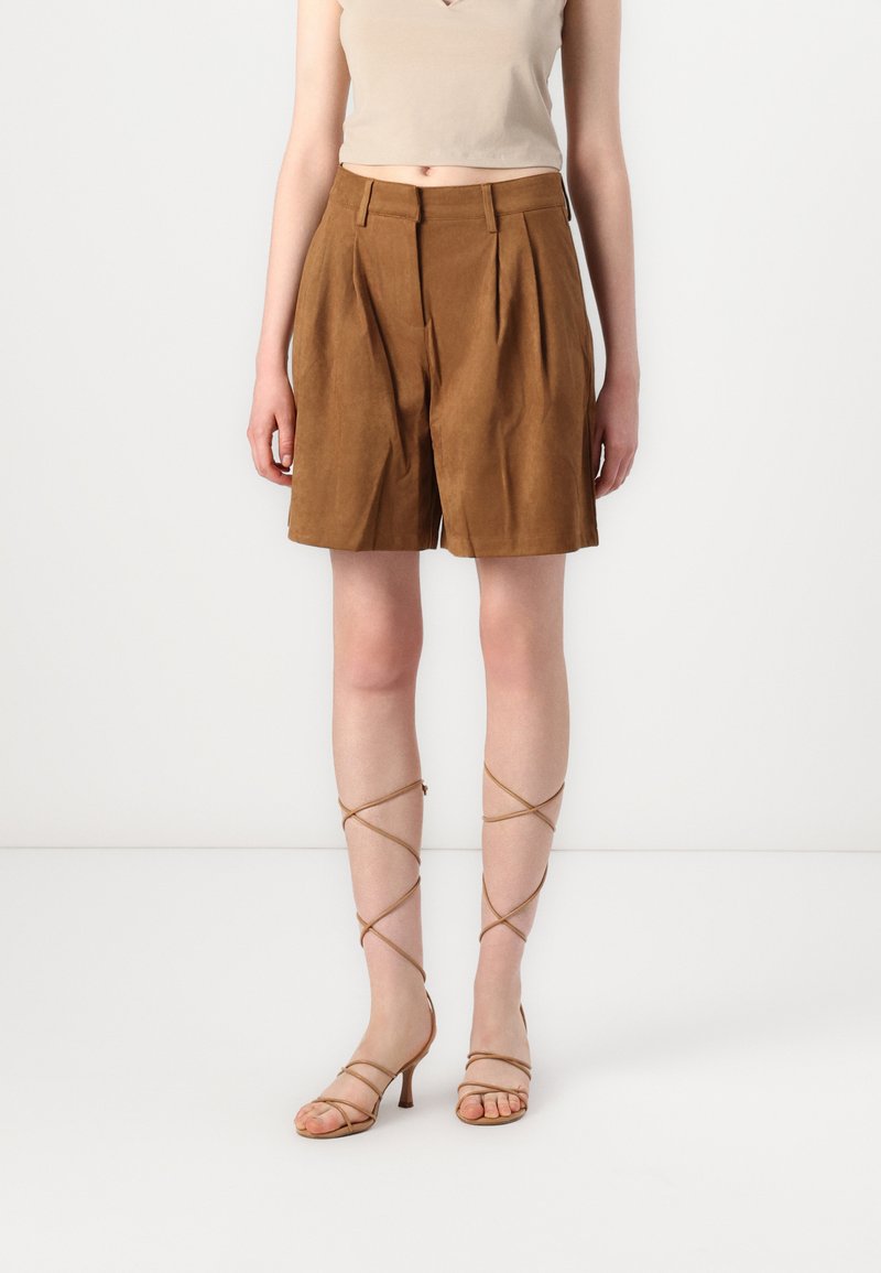 Shorts plissés en daim marron avec une taille mi-haute, associés à des sandales à talons hauts beige avec lanières ; texture lisse et coupe ajustée.