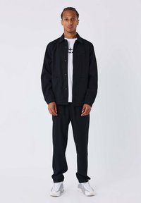 Giacca nera con bottoni, due tasche frontali, sopra una t-shirt bianca, abbinata a pantaloni neri e scarpe da ginnastica grigie. Design semplice, vestibilità casual.
