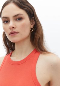 Ripovaný oranžový tílko s kulatým výstřihem. Modelka má dlouhé hnědé vlasy a nosí zlaté kruhové náušnice. Viditelná jemná struktura pleti.