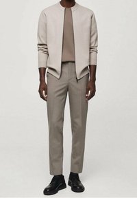 Chaqueta bomber beige claro sobre una blusa topo, combinada con pantalones gris claro y zapatos negros con cordones. Materiales suaves con líneas limpias.